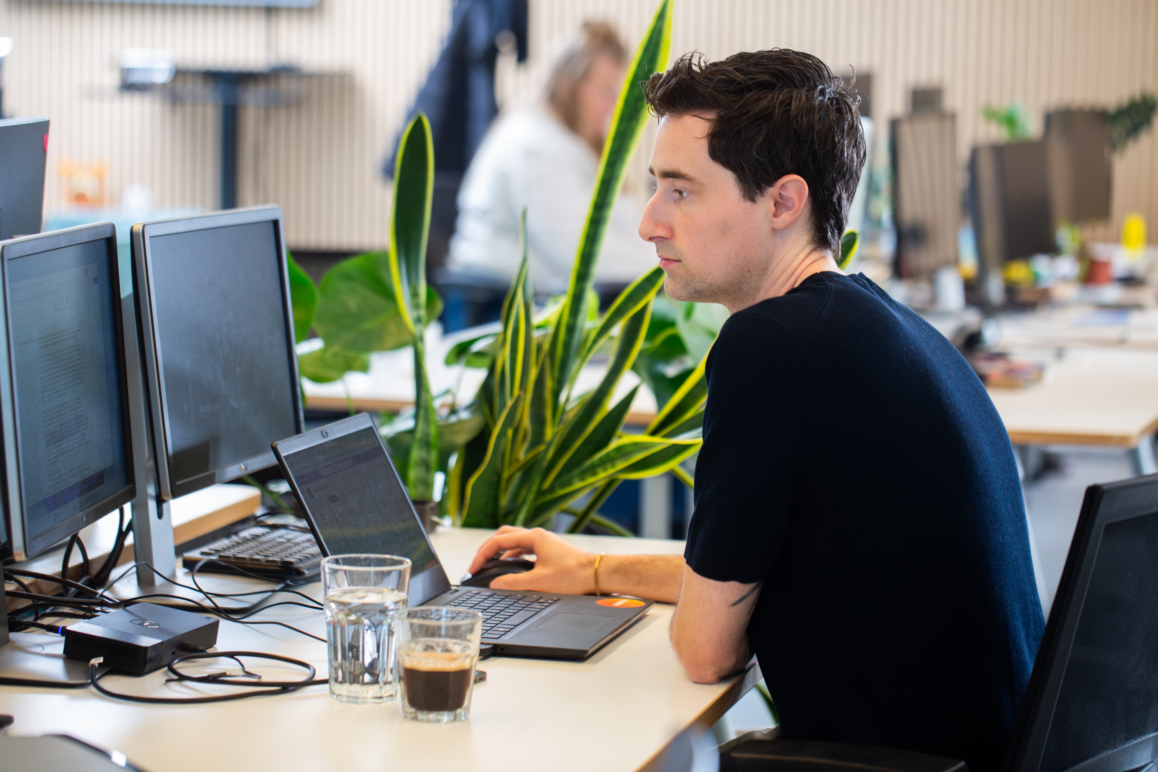 Björn van den Ham werkt geconcentreerd op laptop in modern kantoor met planten en meerdere schermen.